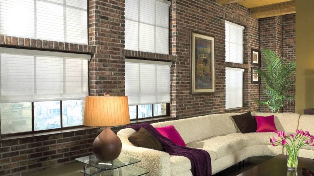 A modern living room with exposed brick walls, large windows with white blinds, a beige sectional sofa with pink and purple pillows, a brown table lamp, wall art, a potted plant, and a vase of pink tulips.